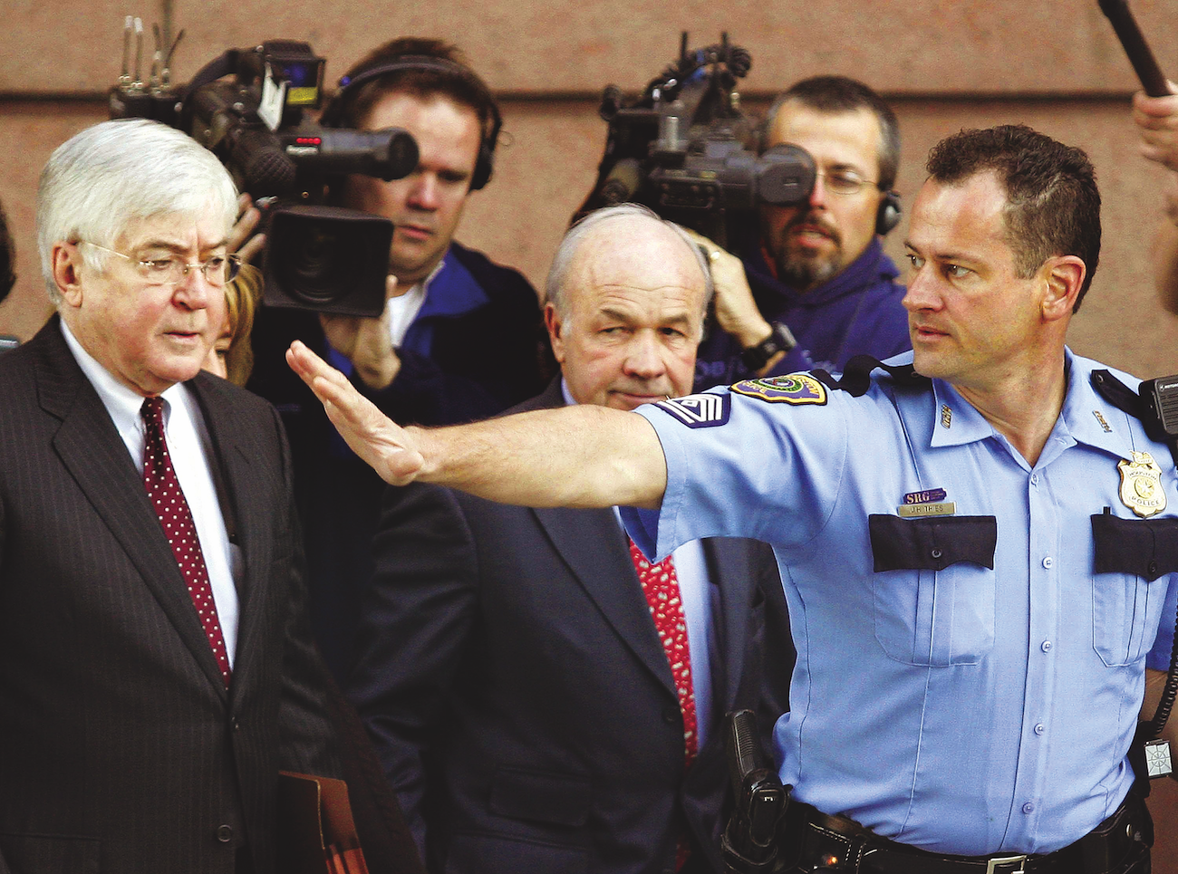 a photograph of a police officer holding his hand in a "stop" motion to a man in a black suit and red tie