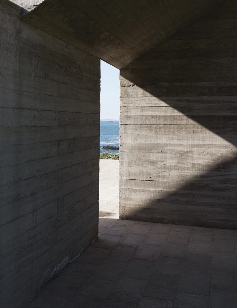 A view of the ocean is framed by the opening between two large, textured concrete walls. Sunlight casts a sharp triangular shadow across the right wall and floor, enhancing the geometric feel of the scene. The blue water and sky provide a serene contrast to the austere, angular architecture.