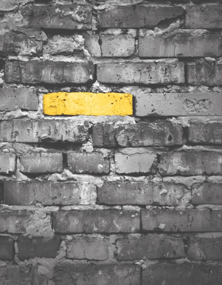 A weathered brick wall is shown in black and white, with the exception of one single horizontal brick that is brightly painted yellow, creating a striking focal point through selective color.