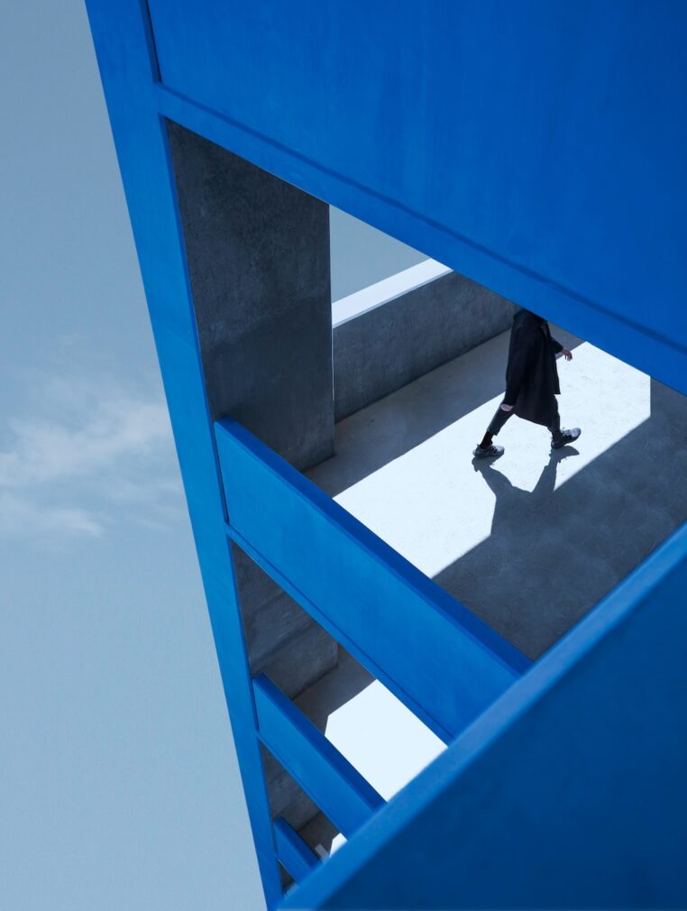 A person walks across a modern architectural structure, framed by bold blue beams and geometric shapes. The composition is angular, with the individual partially obscured, casting a shadow on the sunlit floor. The blue architecture contrasts sharply with the pale sky and concrete surfaces, creating a striking, minimalist visual effect.