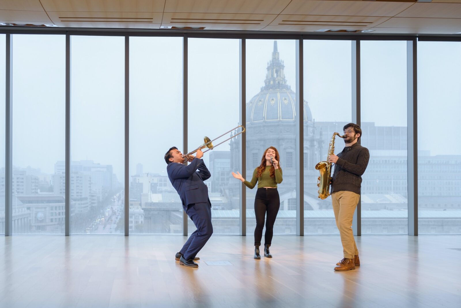 two people playing instruments, the sax and tuba and one person singling in an open studio with glass windows with a gray sky behind them and buildings.