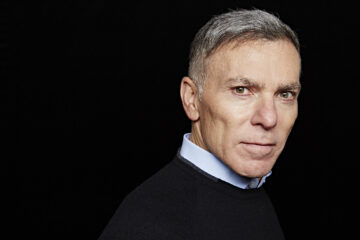 A portrait of Adrian Gore wearing a blue collared shirt and a dark sweater. He is looking confidently at the camera against a plain black background.