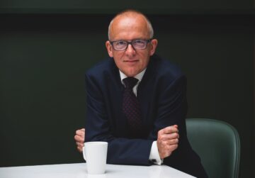Sir Mark Rowley sits at a table with his arms crossed in front of him. He looks directly at the camera with a slight smile. A white coffee mug sits on the table, and the background is plain and dark.