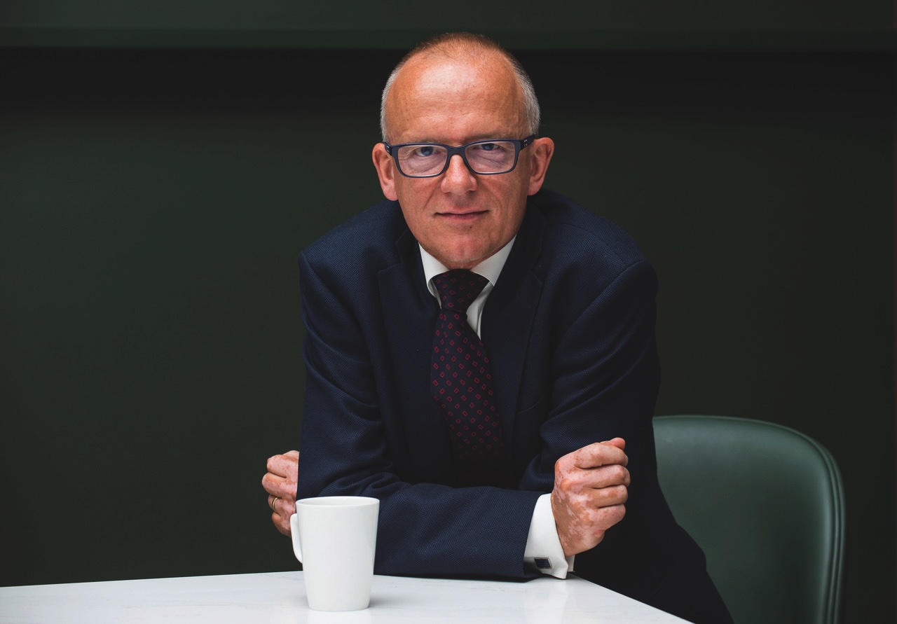 Sir Mark Rowley sits at a table with his arms crossed in front of him. He looks directly at the camera with a slight smile. A white coffee mug sits on the table, and the background is plain and dark.