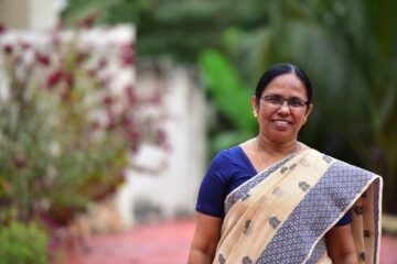 A woman with glasses and a friendly smile stands outdoors, wearing a blue blouse and a cream-colored saree with blue patterns. She has short black hair and is adorned with small gold earrings and a necklace. The background is blurred, featuring greenery and red flowers.