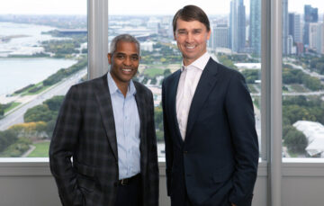 Two professionally dressed men stand side by side, smiling, in front of large windows with a cityscape in the background. One man has short, gray hair and wears a checkered blazer over a light blue shirt. The other has brown hair and wears a classic dark suit with a white shirt. Behind them, tall buildings and water are visible, suggesting an office high above the city.