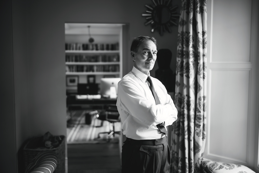 A black-and-white image of Brunswick Group Chairman Sir Alan Parker standing in front of a window with his arms crossed.