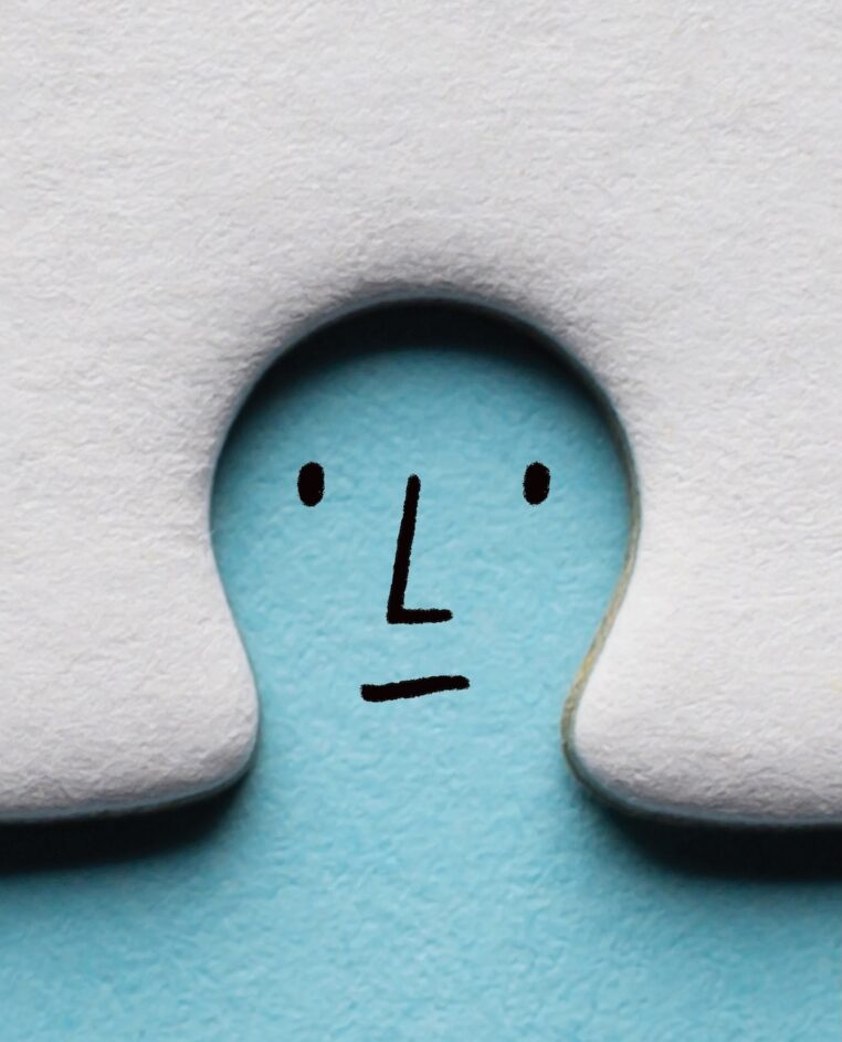 A puzzle piece with a curved cutout reveals a blue background, where a simple, abstract face is drawn with black lines for eyes, a nose, and a mouth. The overall look is minimalist and playful.