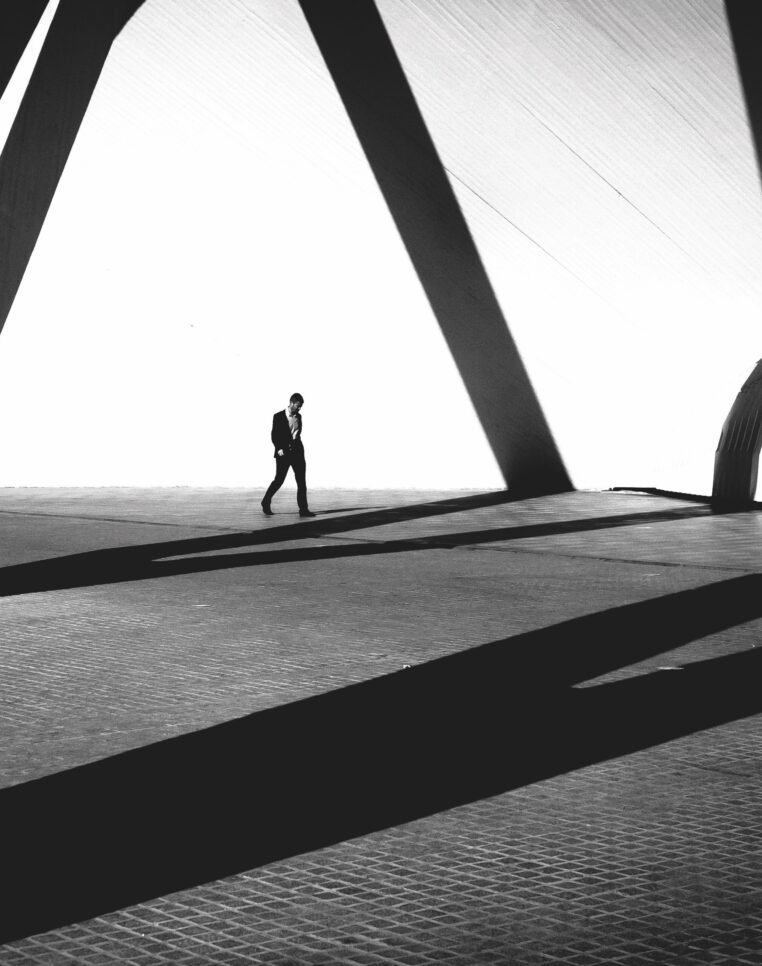 A person in a suit walks alone across a vast, open space with dramatic, geometric shadows cast by large architectural structures. The scene is high-contrast black and white, emphasizing the play between light and shadow and creating a minimalist, modern aesthetic.