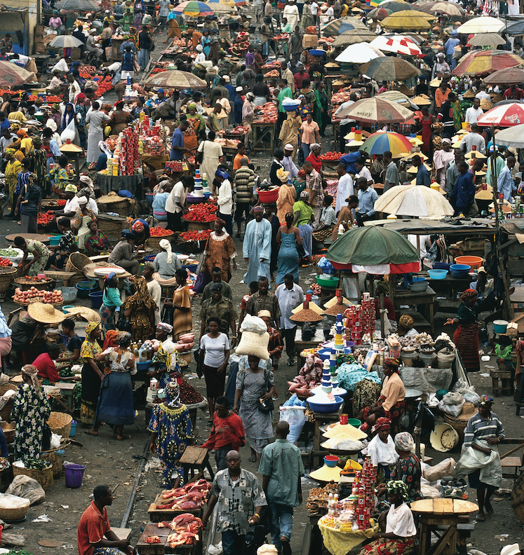 a street crowded with hundreds of people with food and clothing and fabric stands, some with umbrellas over top