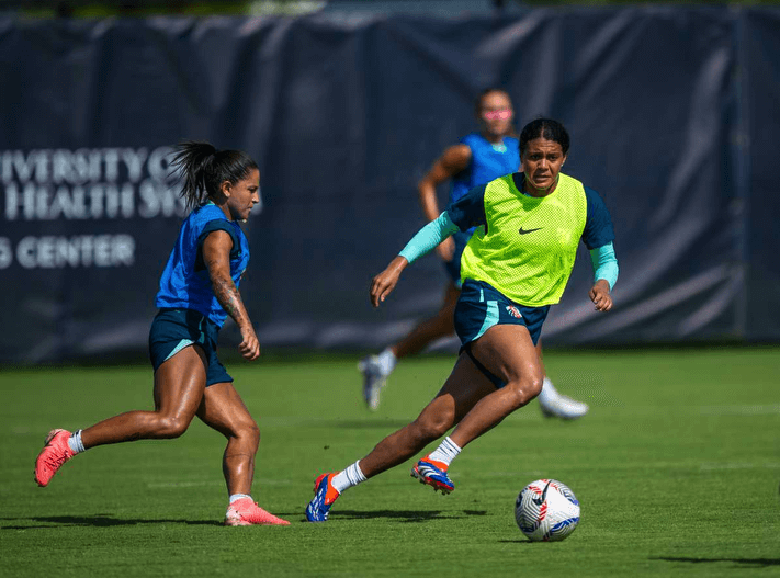 Soccer practice—one player in a neon yellow training bib dribbles while a blue-vested teammate defends on a grass field.