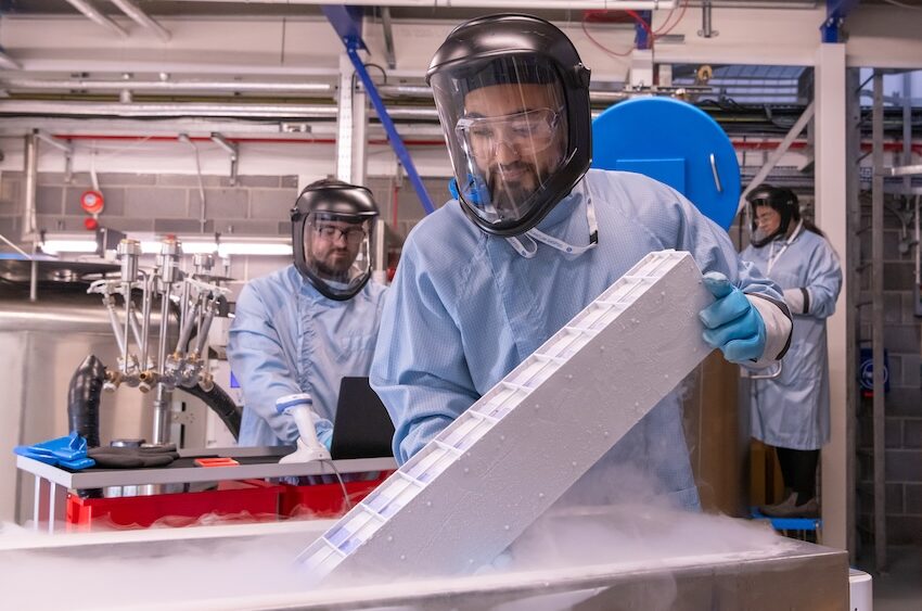 Three scientists with blue medical gowns on and large glass face masks collecting samples in a research lab.