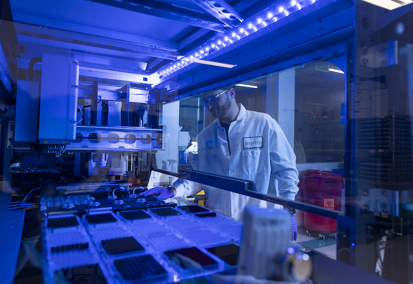 An image of a scientist in a lab coat working with automated equipment and trays under blue lighting in a laboratory.