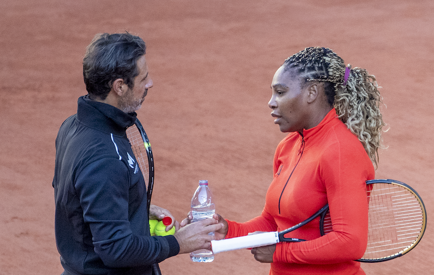 Patrick Mouratoglou and Serena Williams talking on a tennis court handing a bottle of water between them.