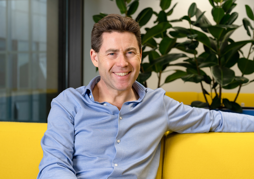 An image of Trustpilot CEO Adrian Blair smiling sitting on a yellow couch with a plant behind him.