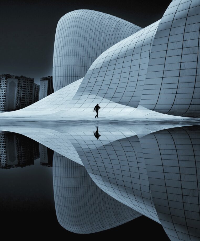 A person walks across a smooth, futuristic architectural landscape with flowing, curved structures covered in a grid pattern. The scene is reflected perfectly below, creating a symmetrical, almost surreal effect. Tall buildings are visible in the background, adding contrast to the modern foreground design. The overall tone is cool and monochromatic, emphasizing the dramatic shapes and the solitary figure.