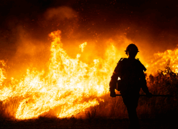 A firefighter is silhouetted against a raging wildfire at night. Massive flames and thick smoke fill the background, illuminating the firefighter's protective gear as they stand on the edge of the burning area, holding equipment. The scene is intense and dramatic, highlighting the danger and urgency of wildland firefighting.