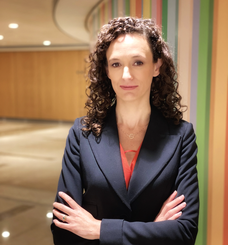 A woman with curtly brown hair in a navy suit jacket stands with her arms crossed against a colorful wall.