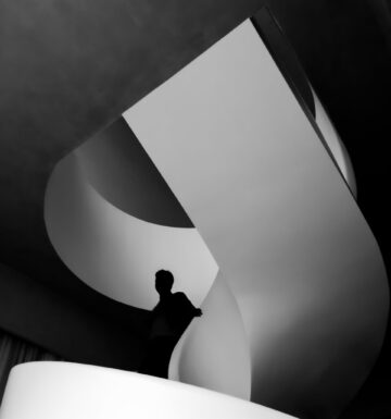 A black and white photograph of a person standing on a modern, spiral staircase. The image emphasizes dramatic light and shadow, with the silhouette of the person contrasting against the smooth, curving lines of the staircase. The composition creates a sense of abstraction and architectural elegance.
