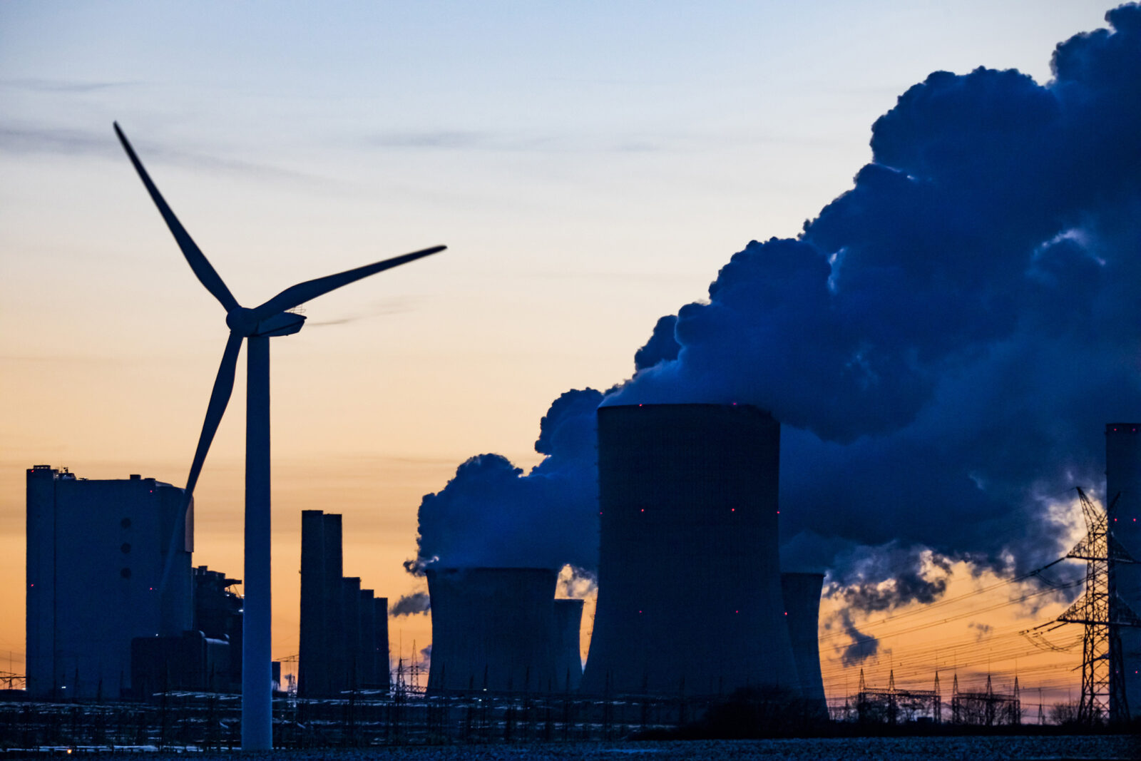 Silhouette of a wind turbine beside a coal power plant with cooling towers emitting smoke at sunset.