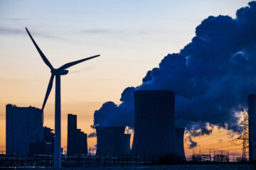 Silhouette of a wind turbine beside a coal power plant with cooling towers emitting smoke at sunset.