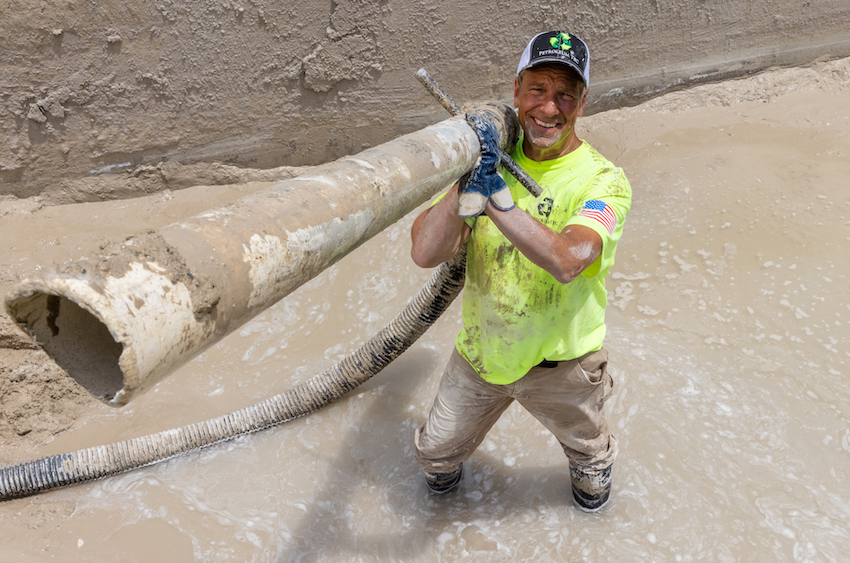 Mike Rowe standing in a foot of muddy water holding a muddy hose smiling for the camera