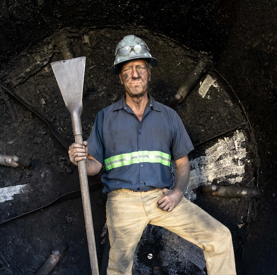 Mike Rowe standing in front of a metal mine entrance covered in black dirt wearing a helmet and holding a metal shovel