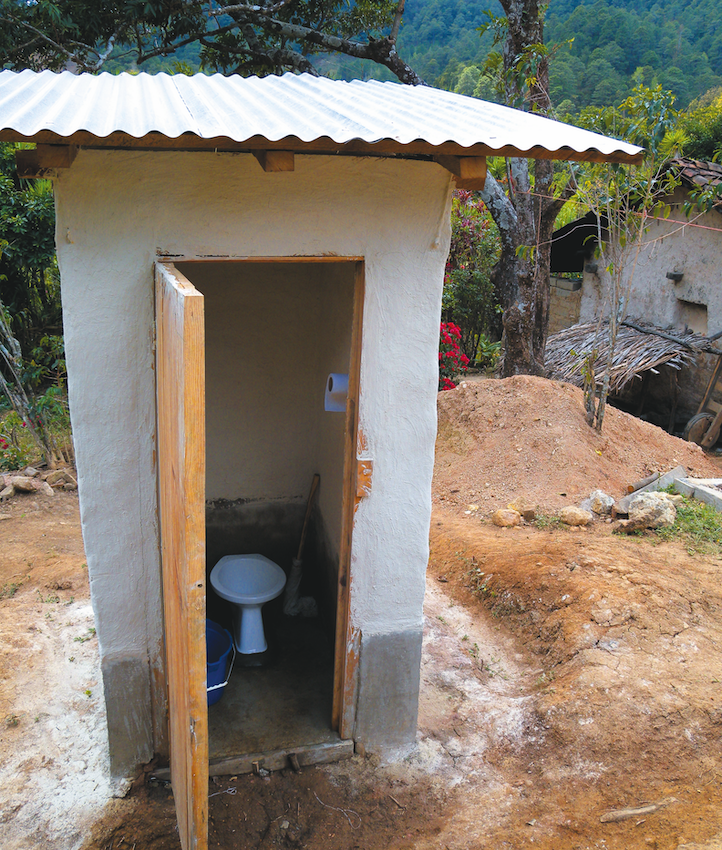 Small outdoor latrine with a corrugated metal roof and an open door showing a toilet inside, set in a rural area with trees and mountains in the background.