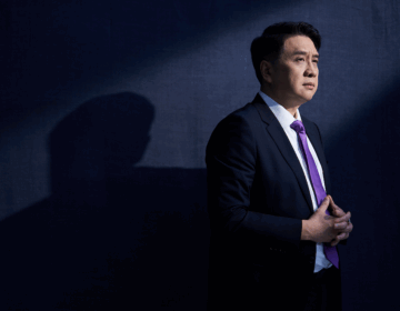 Man in a dark suit with a purple tie standing against a dark background, hands clasped, looking thoughtful with light casting a shadow behind him.