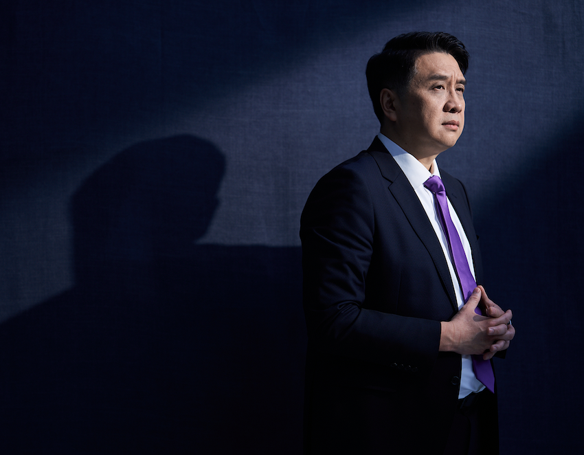 Man in a dark suit with a purple tie standing against a dark background, hands clasped, looking thoughtful with light casting a shadow behind him.