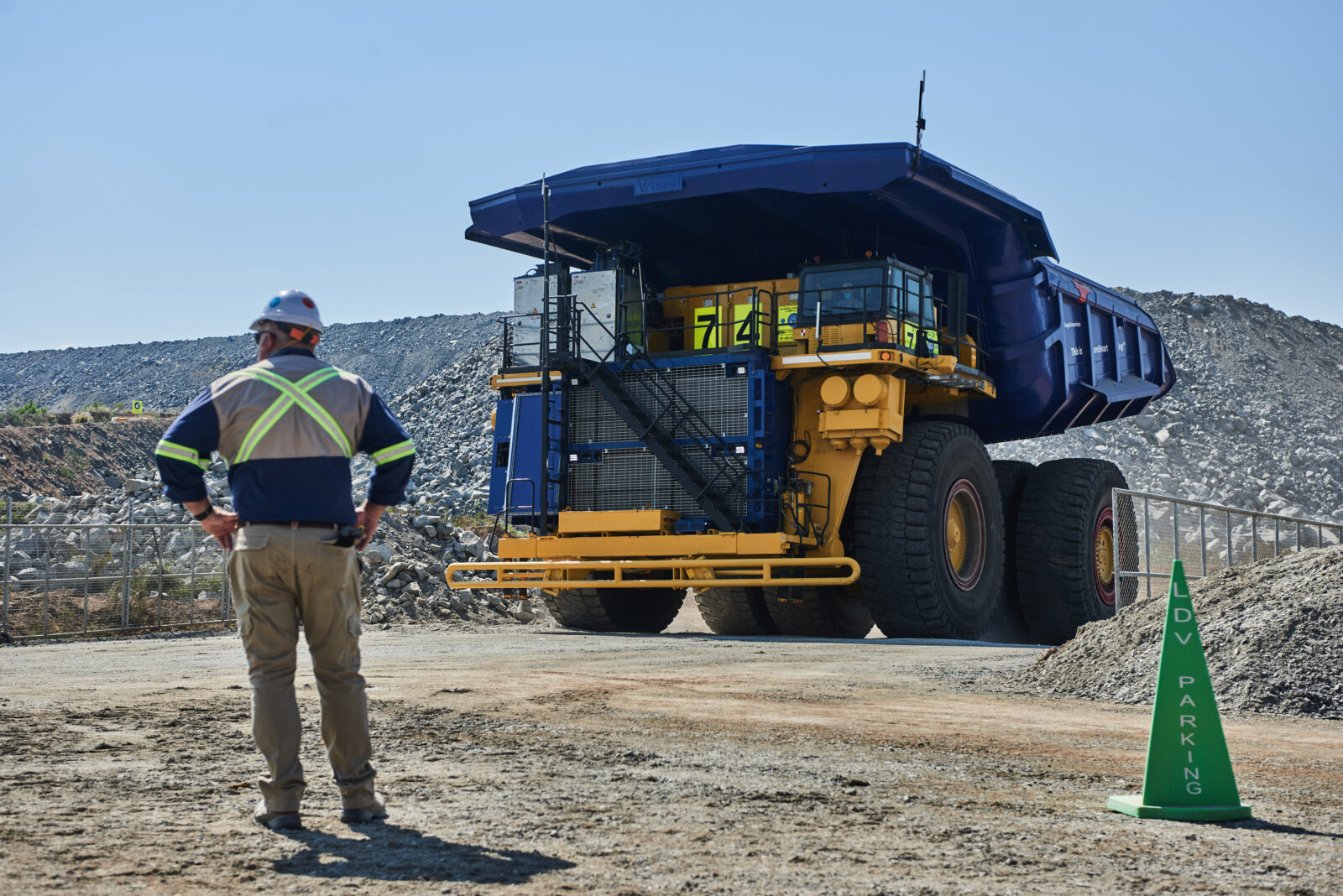 Worker in safety gear stands in front of a massive yellow and blue mining dump truck at a quarry site, with rocky terrain in the background.