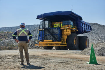 Worker in safety gear stands in front of a massive yellow and blue mining dump truck at a quarry site, with rocky terrain in the background.