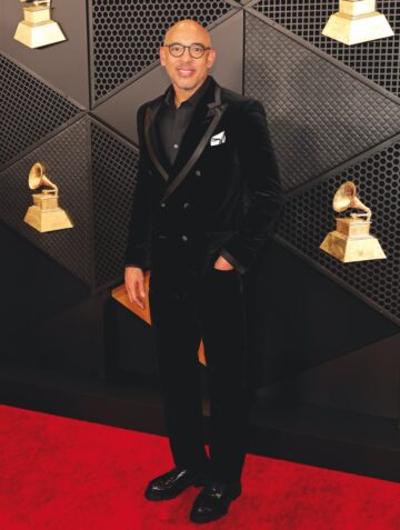 A man in a black tux with a black shirt on a red carpet with the gold Grammy Award on the black step & repeat behind him.