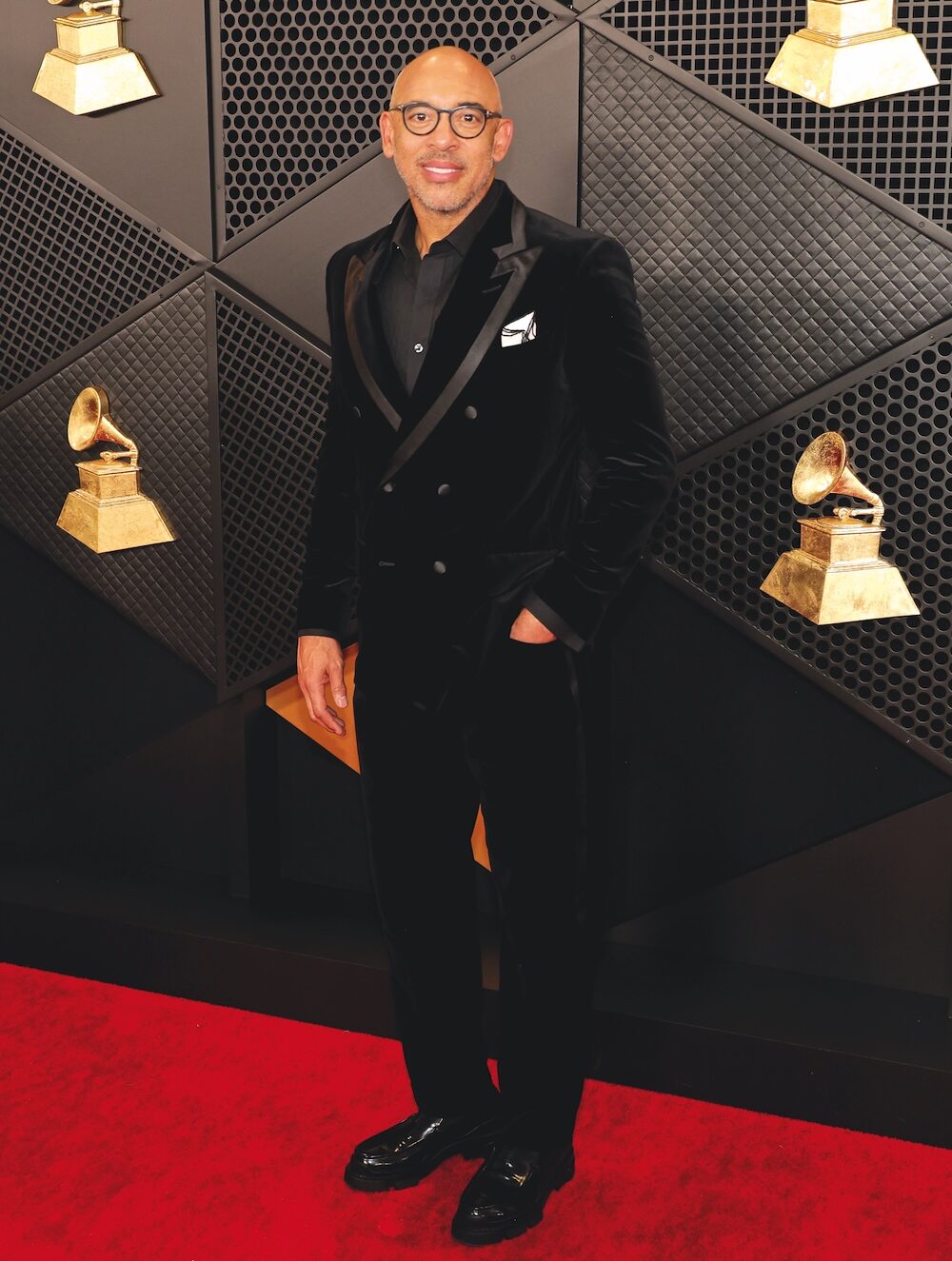 A man in a black tux with a black shirt on a red carpet with the gold Grammy Award on the black step & repeat behind him.