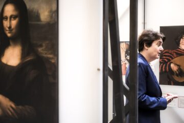 Person in a blue suit speaking inside an art gallery, with Leonardo da Vinci’s Mona Lisa painting visible on the left and other artworks in the background.