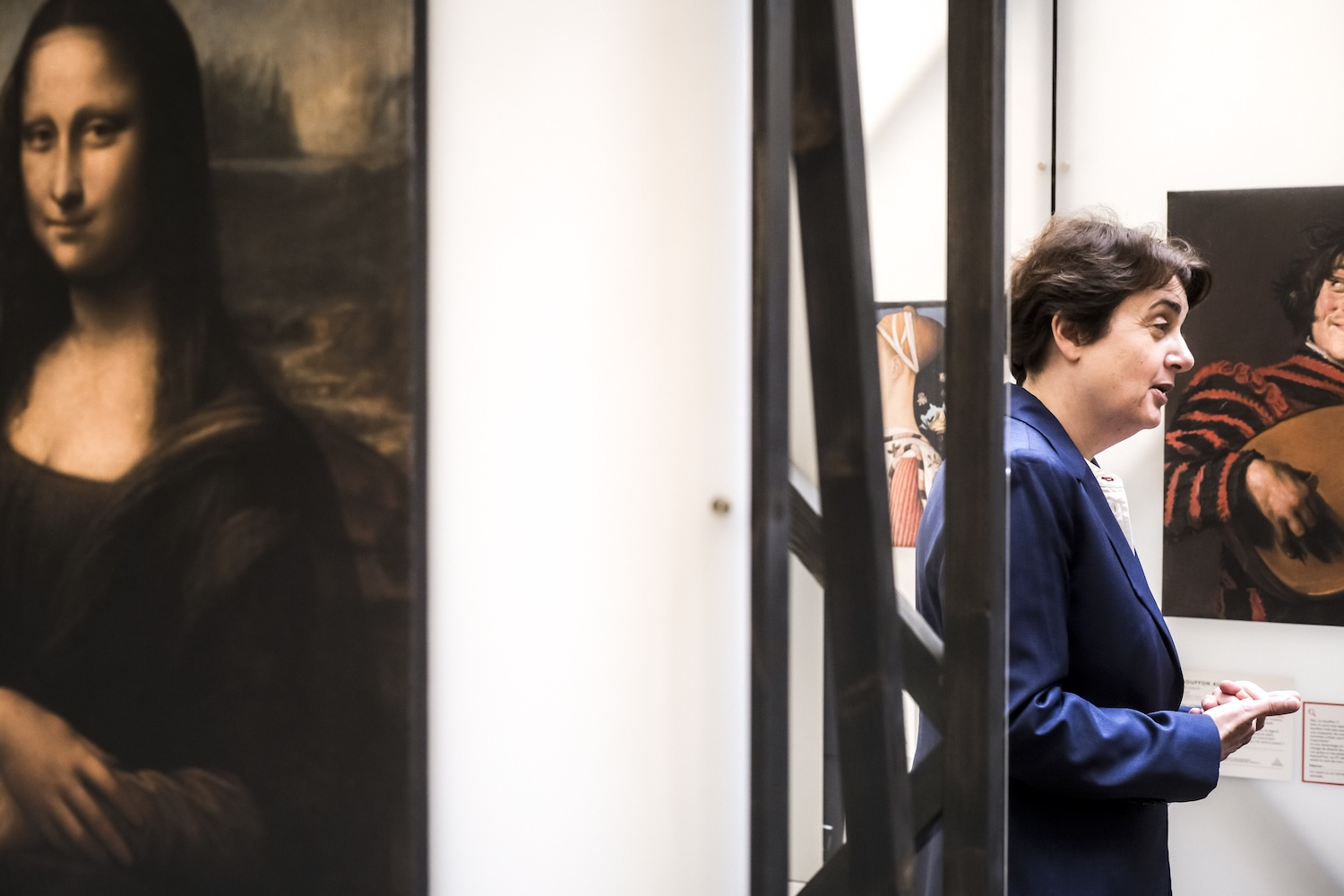 Person in a blue suit speaking inside an art gallery, with Leonardo da Vinci’s Mona Lisa painting visible on the left and other artworks in the background.