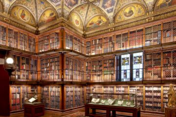 Ornate historic library interior with two levels of bookshelves enclosed by decorative metal grilles, stained glass windows, illuminated ceiling murals, and display cases with open books.