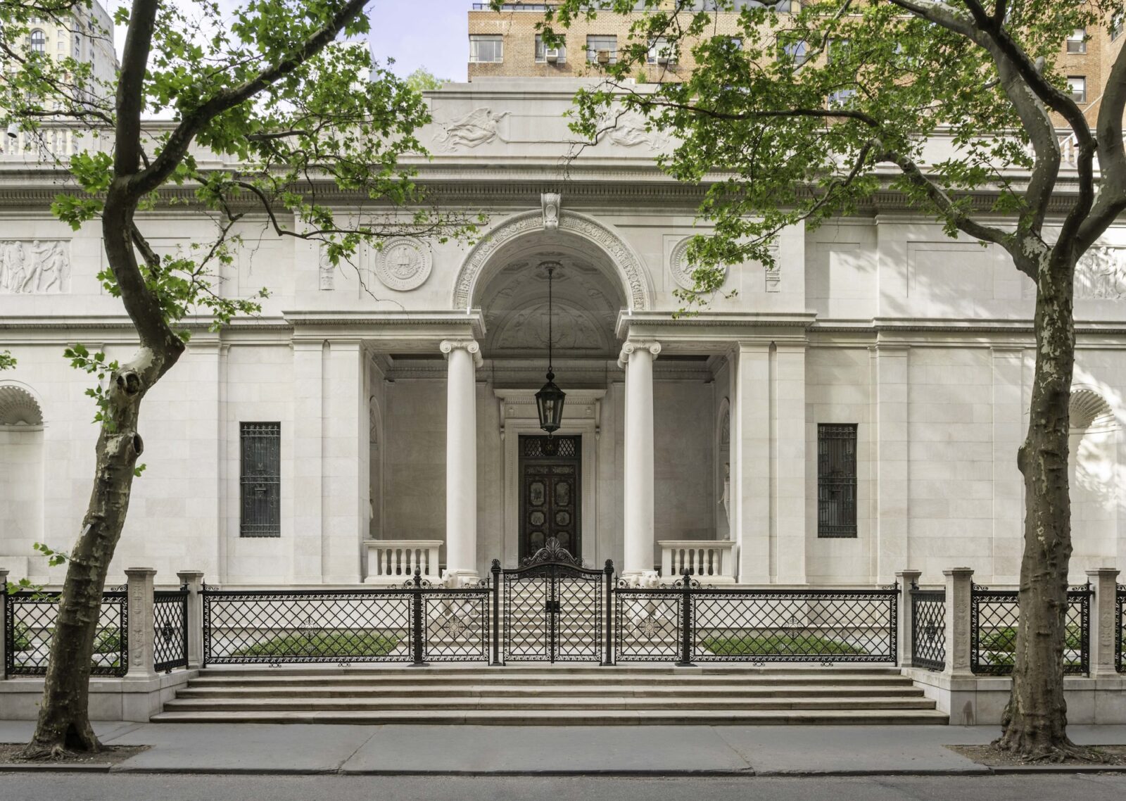 The front of the Morgan Library, a cream colored building with a large archway, framed by two trees