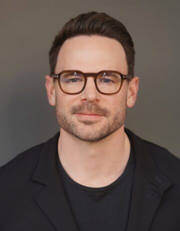 A headshot of a man with glasses and brown hair wearing a black shirt and black suit jacket.