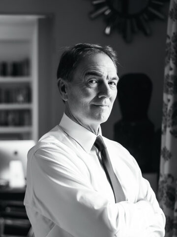 A black-and-white image of Brunswick Group Chairman Sir Alan Parker standing in front of a window with his arms crossed.