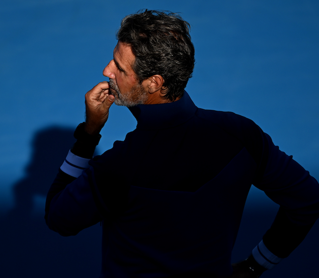 Man with salt-and-pepper hair standing against a blue background, looking thoughtful with his hand on his chin.