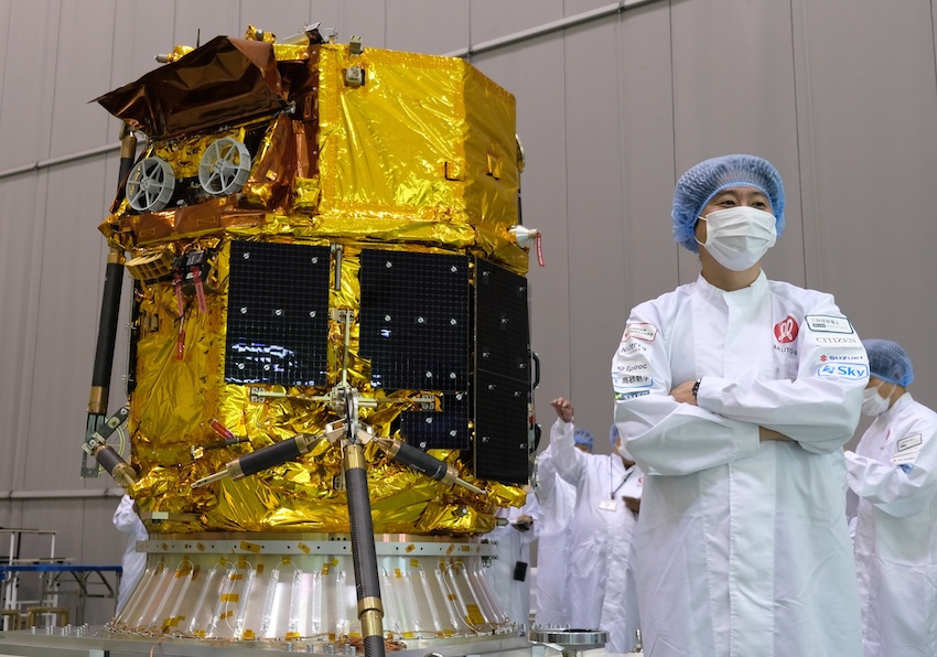 A man in a white lab coat, paper face mask, and blue hair net stands next to a gold robotic lunar lander.