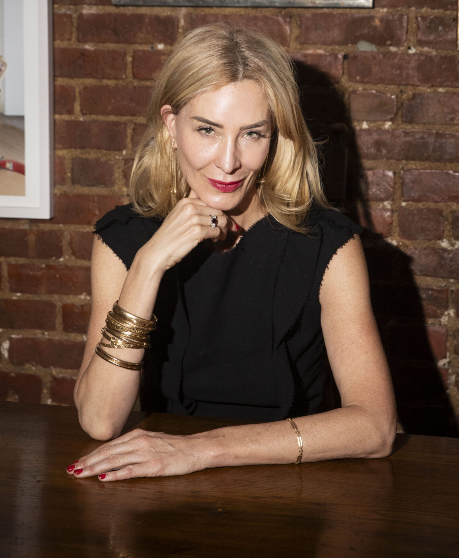 Blonde woman wearing a black dress sitting at a table in front of a brick wall with her hand under her chin