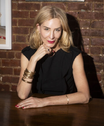 Blonde woman wearing a black dress sitting at a table in front of a brick wall with her hand under her chin