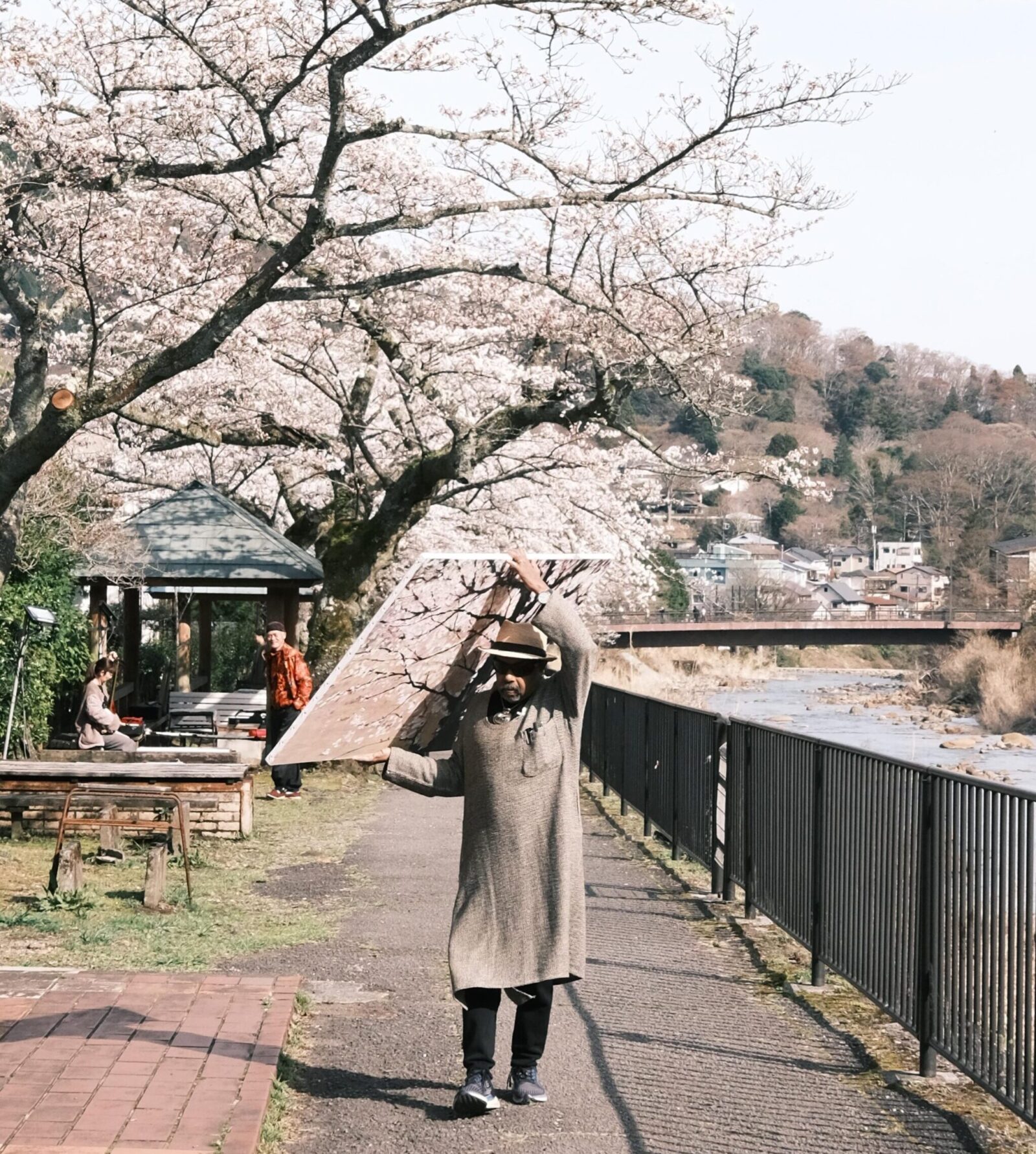 A person wearing a long coat and hat carries a large reflective board under blooming cherry blossom trees along a riverside path. Other people are seated and standing nearby under a small gazebo, with more cherry trees and a bridge in the background.
