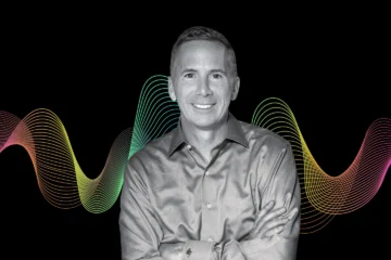 black and white photograph of a man smiling in a dress shirt with her arms crossed in front of his chest. Behind him a black background with orange, yellow, green soundwave graphics.
