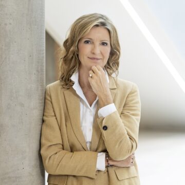 image of a blonde woman in a tan suit with a white button down leaning against a concrete pole with her hand resting against her chin