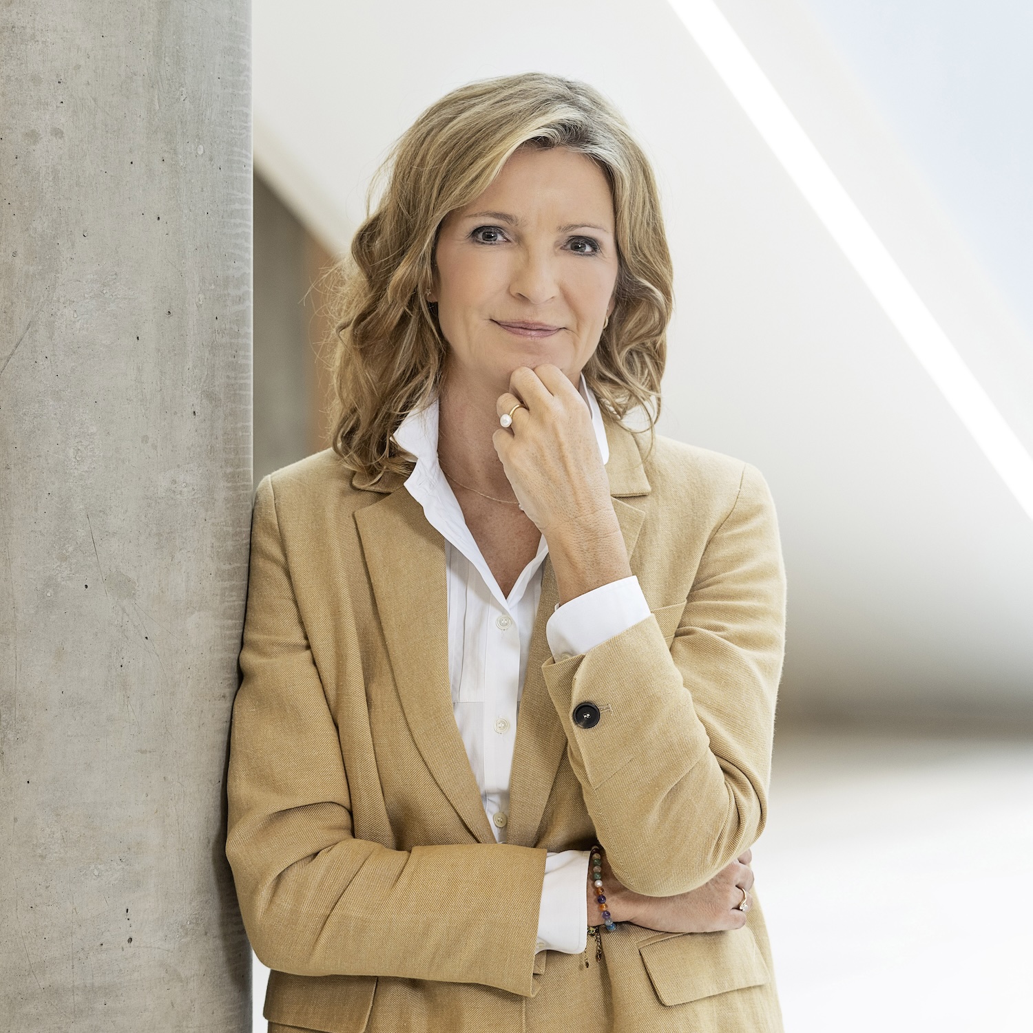 image of a blonde woman in a tan suit with a white button down leaning against a concrete pole with her hand resting against her chin