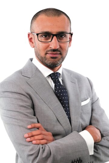 headshot photograph of a man in black rimmed glasses and a light gray suit with a black tie and white polka dots. His arms are crossed across his chest.