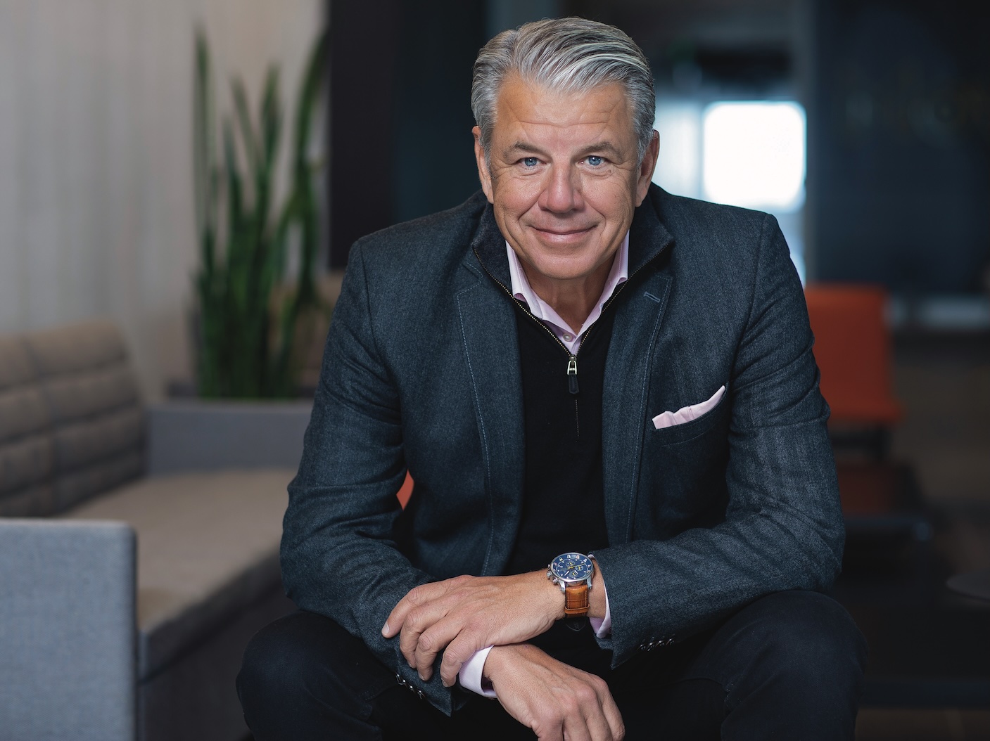 a man with gray hair in a navy suit jacket over a sweater sitting on a chair leaning over resting his forearms on his knees and smiling